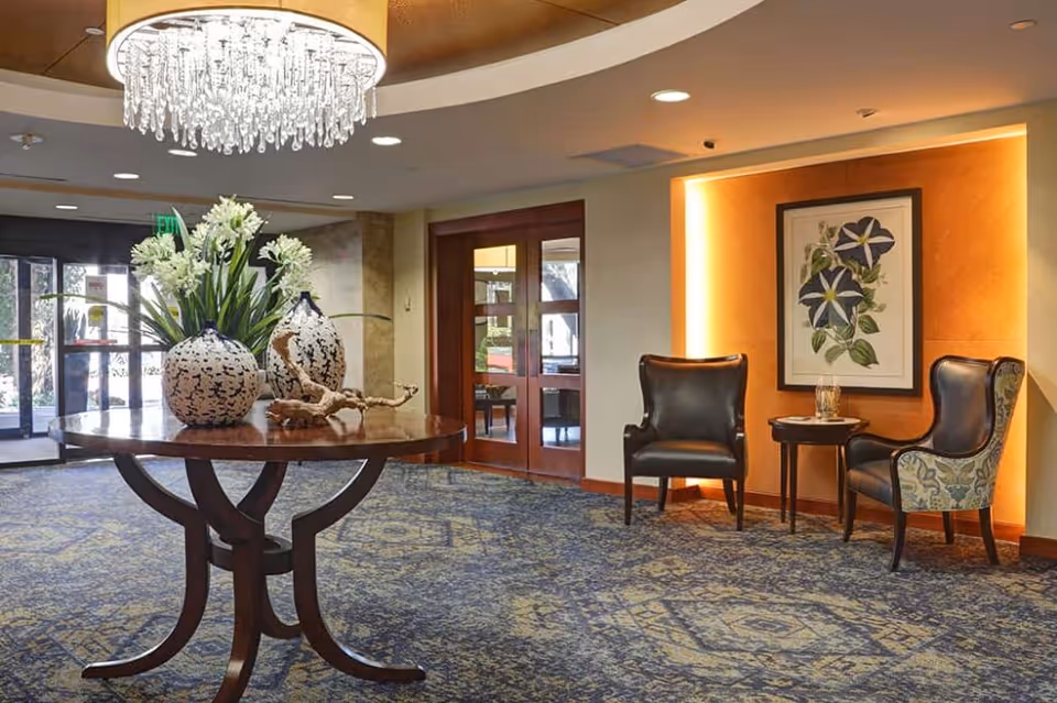 Lobby-style interior with a round table topped with vases and flowers, a chandelier overhead, and two chairs against an orange-accented wall.