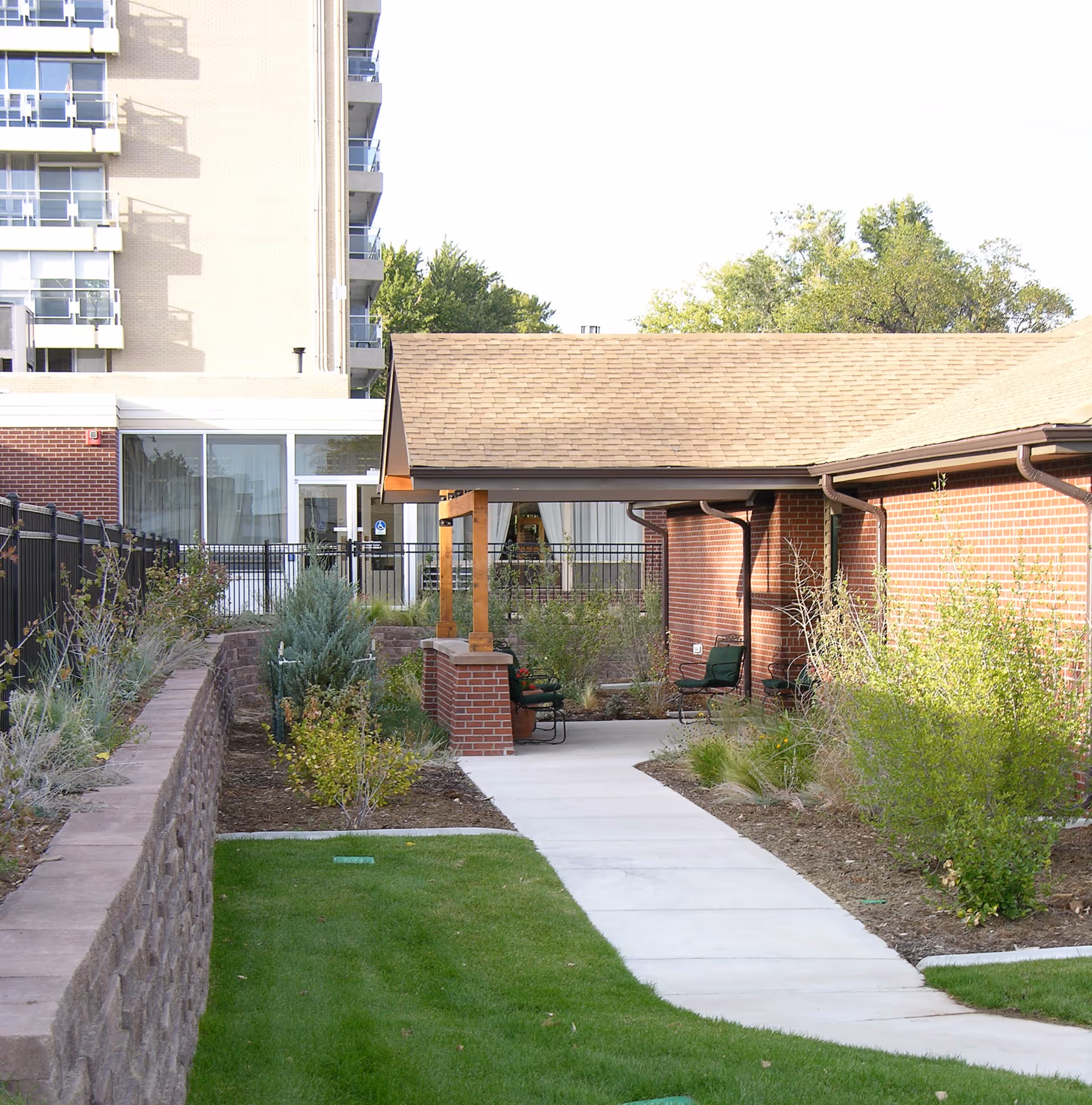 Outdoor view of a senior living facility showing a paved walkway leading to a covered seating area with chairs. The area is surrounded by well-maintained landscaping including grass, shrubs, and small trees. The building has red brick walls and a tan shingled roof. In the background, there is a taller building with balconies and large windows.
