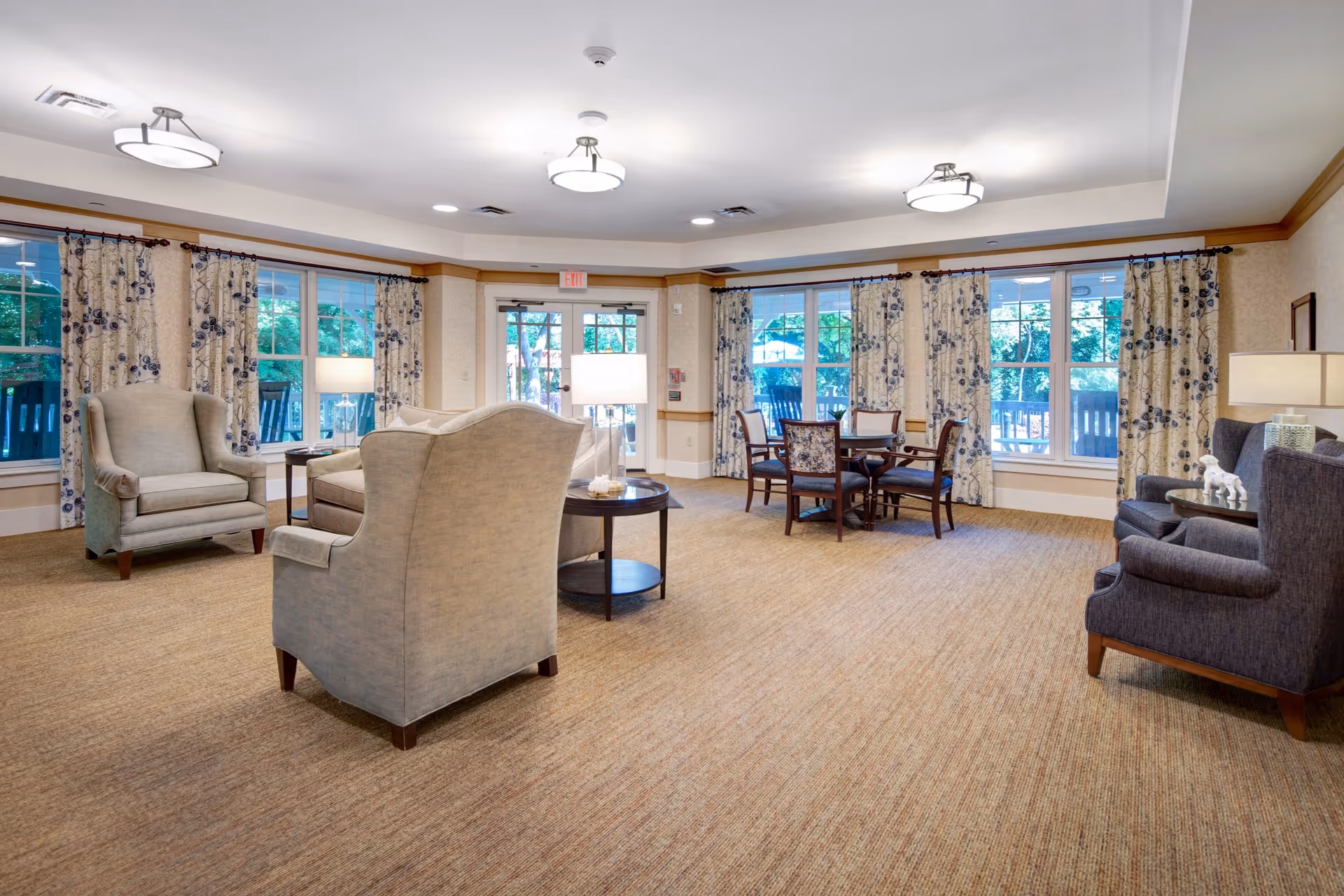 A spacious, well-lit common area with large windows covered by floral curtains. The room features comfortable armchairs, a round table with chairs, side tables with lamps, and beige carpeting. The walls are light-colored with wooden trim, and ceiling lights provide additional illumination.