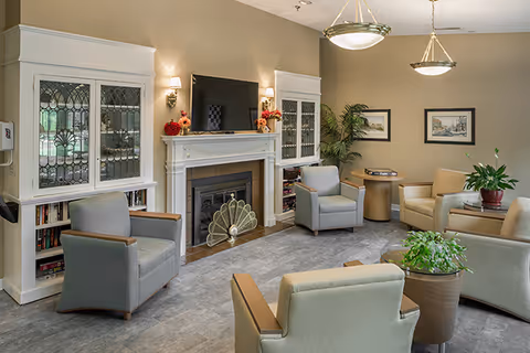 Sunlit common living room with armchairs arranged around a fireplace and television, built-in cabinets, plants, and framed artwork.