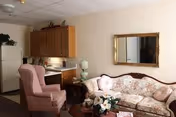 Small senior living room with a floral sofa, pink armchair, coffee table, and a kitchenette with wooden cabinets and refrigerator.