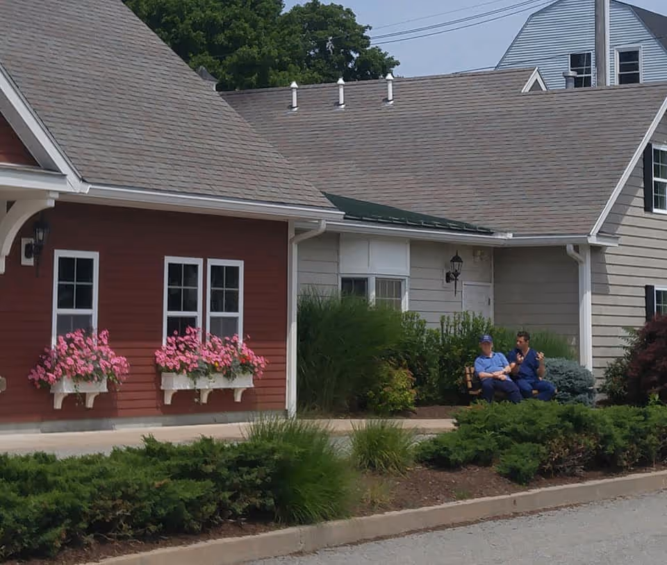 Exterior view of a senior living facility named The Elms, showing a red building with white-trimmed windows adorned with flower boxes filled with pink flowers. Two people are sitting and talking on a bench near the building, surrounded by green bushes and landscaping.