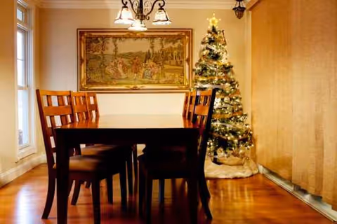 A wooden dining table with chairs in a warm dining room, Christmas tree nearby.