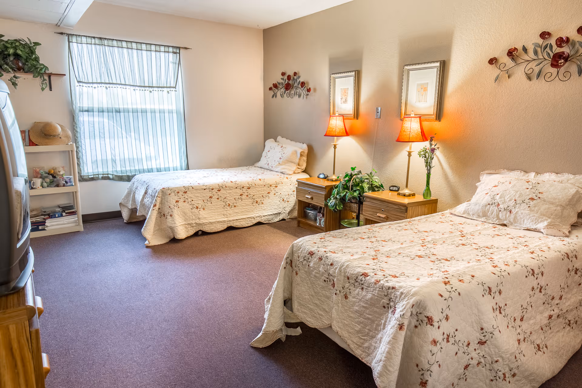 A cozy bedroom with two twin beds covered in white floral bedspreads. Between the beds are two wooden nightstands with table lamps, a small plant, and decorative items. The room has beige walls with floral wall decorations and two framed pictures. A window with sheer striped curtains lets in natural light. A small white shelf with books, a hat, and stuffed animals is visible near the window. The floor is carpeted in a muted purple color.