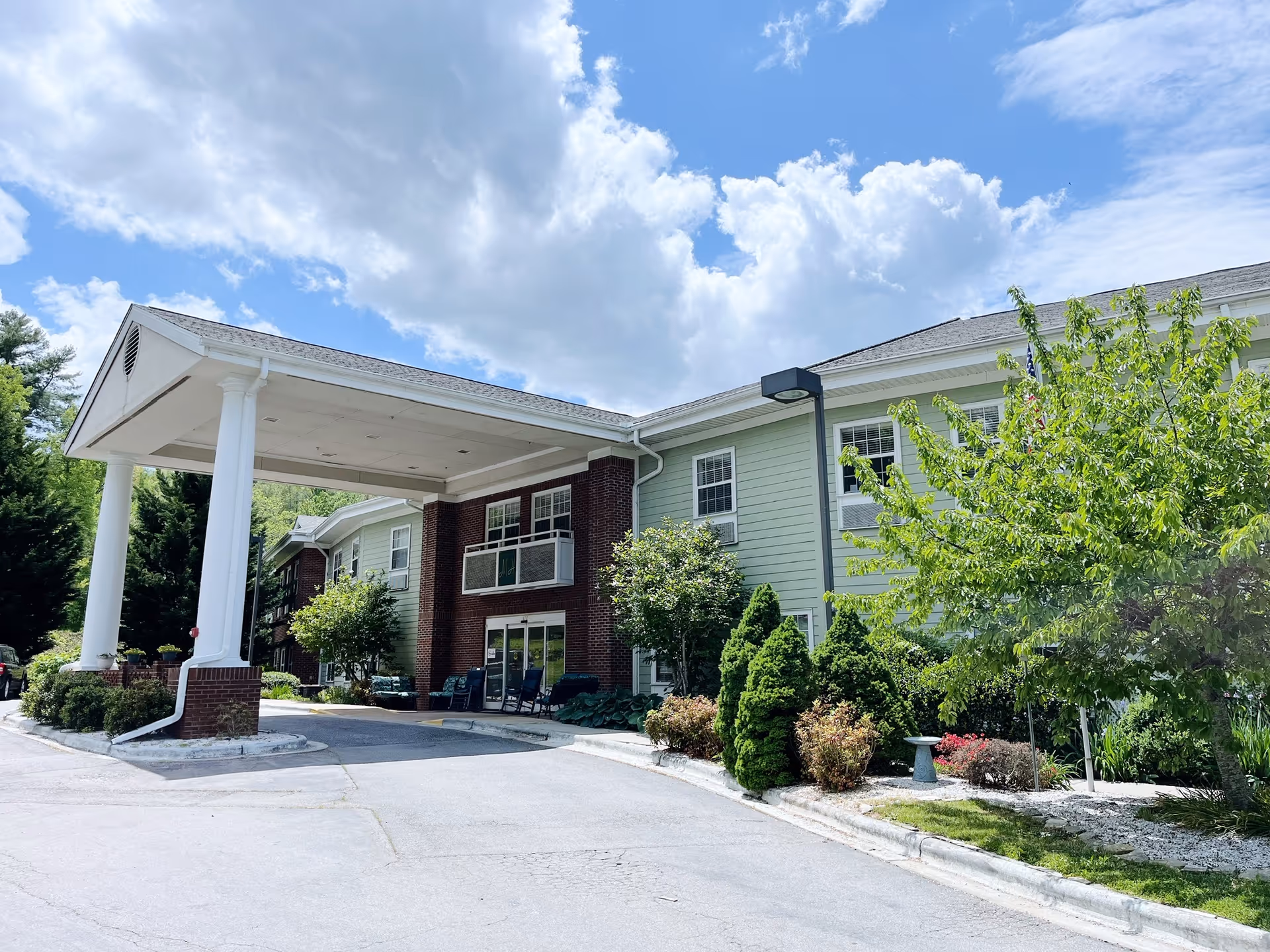 Exterior view of Mars Hill Manor, a two-story building with a covered entrance supported by white columns. The building has a combination of brick and light green siding, surrounded by well-maintained landscaping including trees and shrubs. The sky is partly cloudy with blue patches visible.