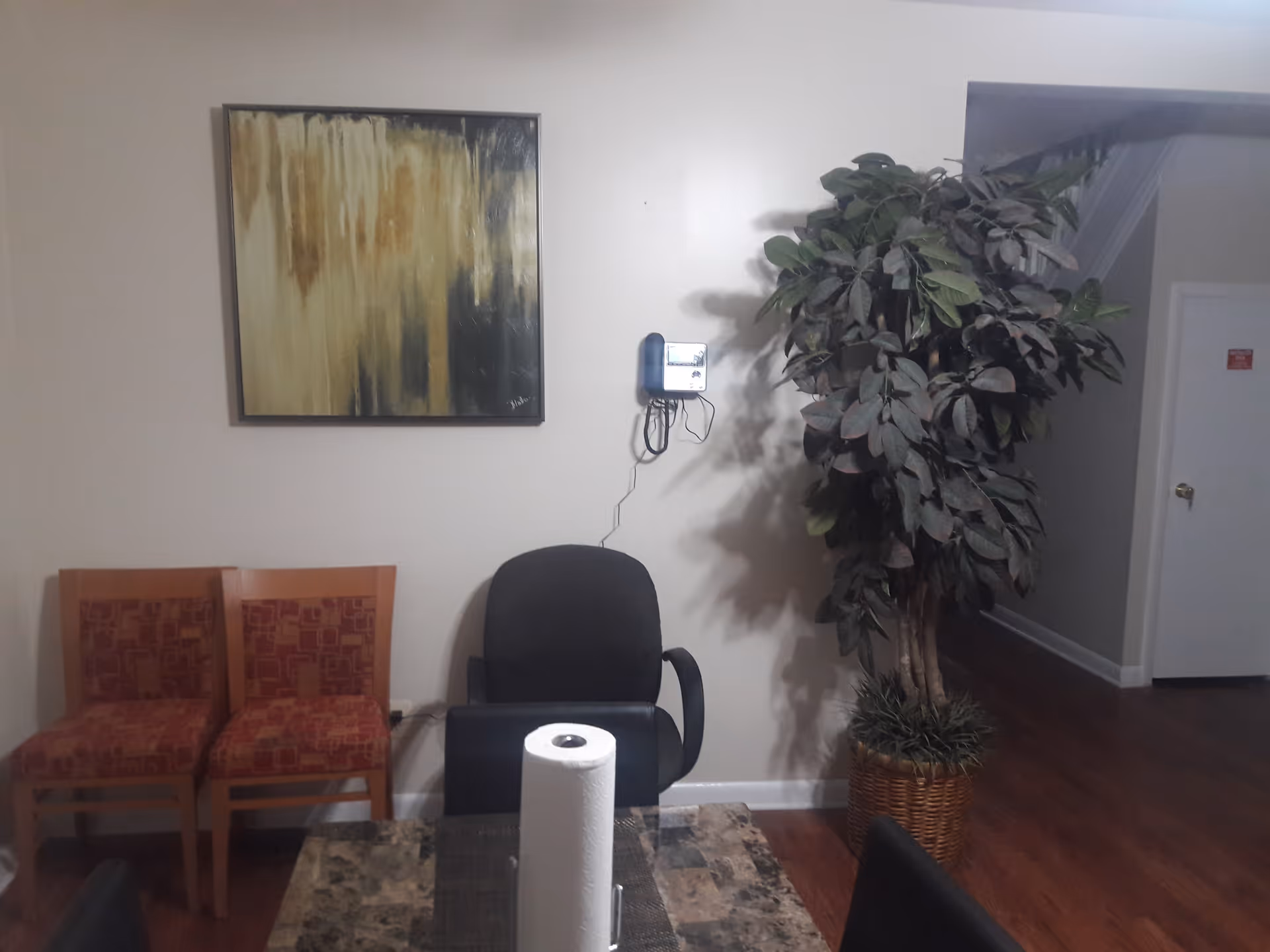 Interior view of a room with a table in the foreground holding a roll of paper towels, two wooden chairs with red patterned cushions against a wall, a black office chair, a large potted plant, a wall-mounted telephone, and an abstract painting on the wall.