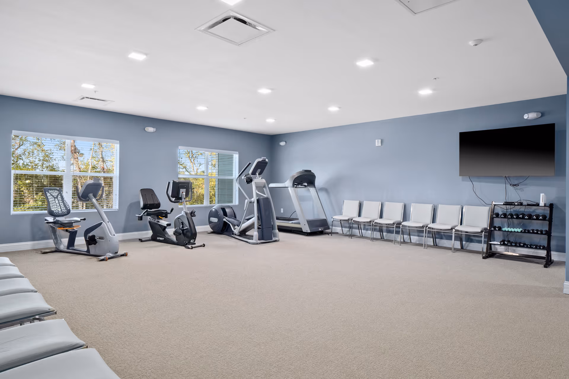 Bright fitness room with exercise machines, a row of chairs, a wall-mounted TV and a dumbbell rack.