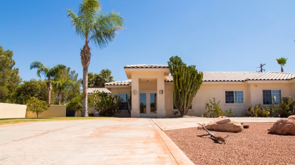 Exterior view of a single-story building with a tiled roof, desert landscaping including cacti and rocks, palm trees, and a clear blue sky.