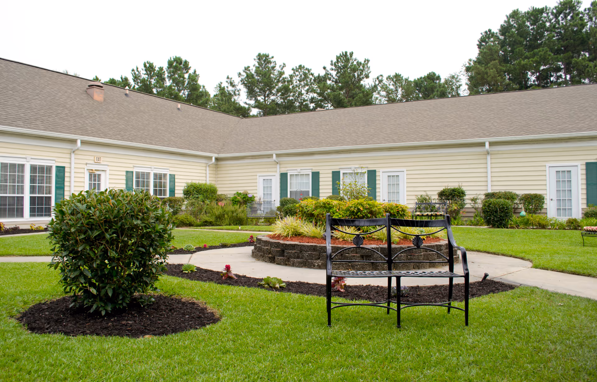 Outdoor courtyard area at The Legacy of Hartsville featuring a circular garden bed with shrubs and plants, surrounded by a paved walkway. There is a black metal bench in the foreground on a well-maintained green lawn. The building with beige siding and green shutters encloses the courtyard, with several windows and doors visible. Trees are seen in the background beyond the building.