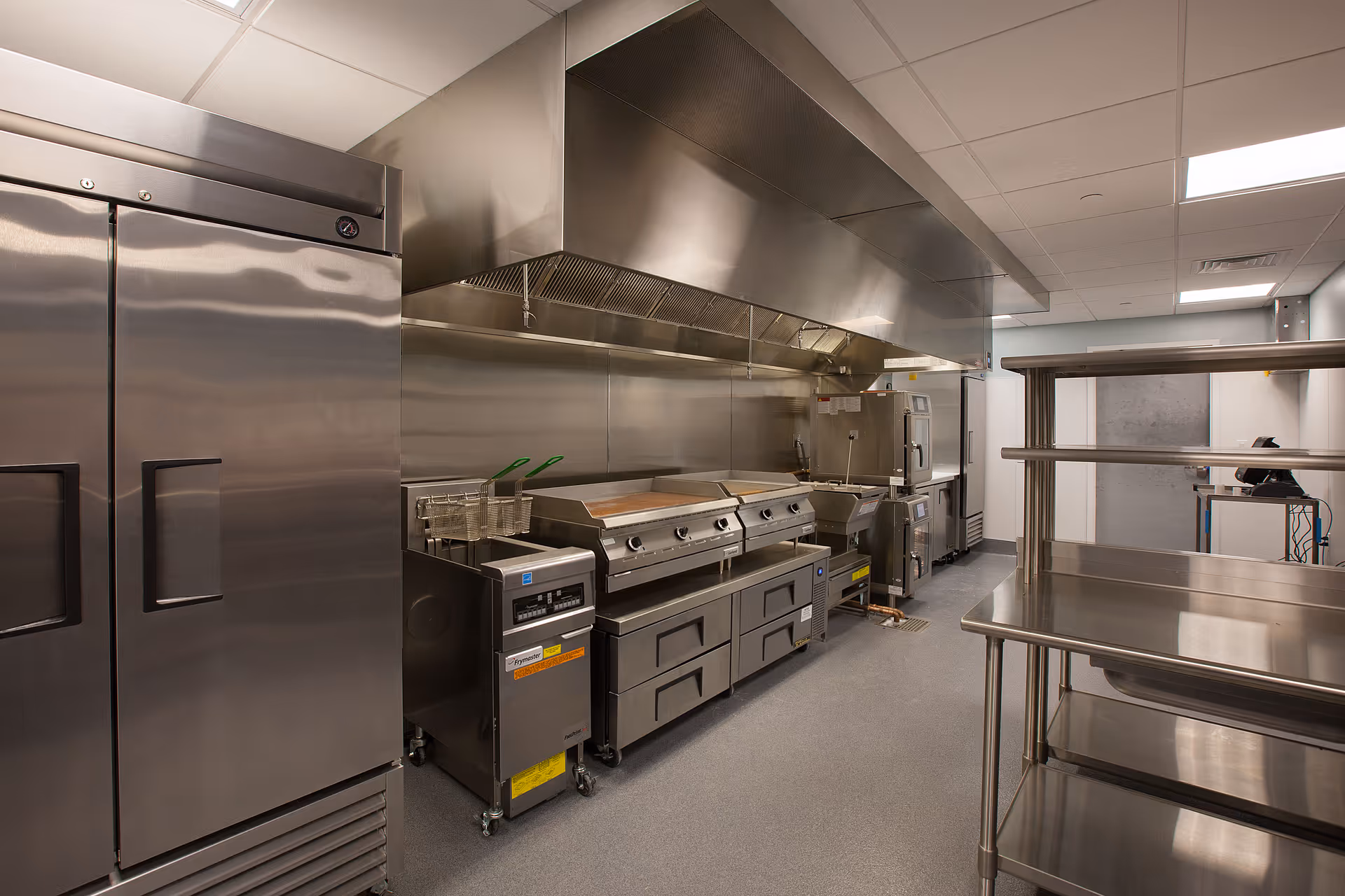A clean, modern commercial kitchen with stainless steel appliances including a large refrigerator, a flat-top grill with ventilation hood, fryers, and metal shelving units. The kitchen has a gray floor and white ceiling tiles with fluorescent lighting.