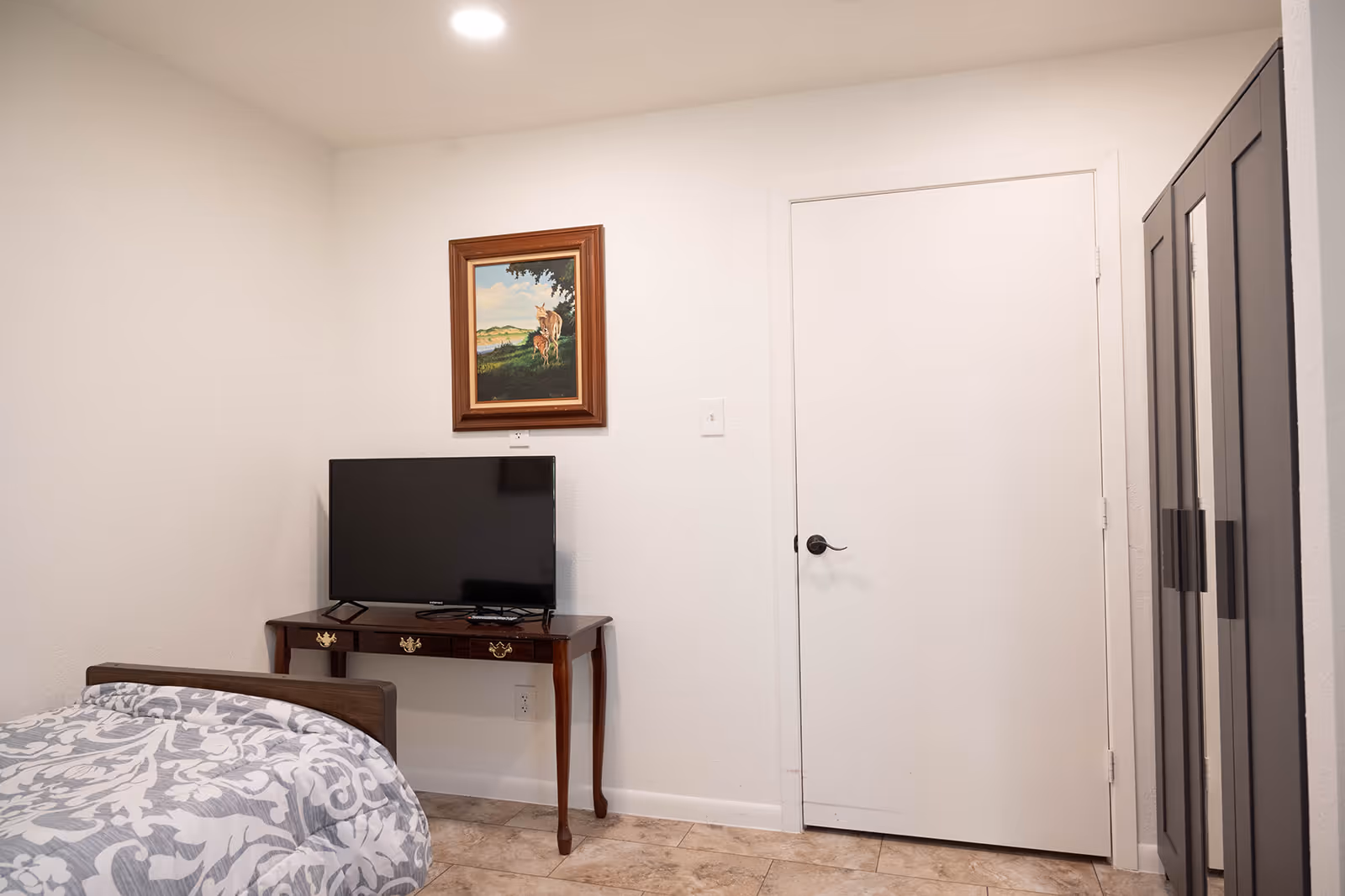 A simple bedroom with a bed partially visible on the left covered with a patterned gray and white blanket. A wooden table against the wall holds a flat-screen TV. Above the TV hangs a framed painting of a deer in a natural landscape. To the right is a closed white door and a dark-colored wardrobe with mirrored doors. The floor is tiled and the walls are painted white.