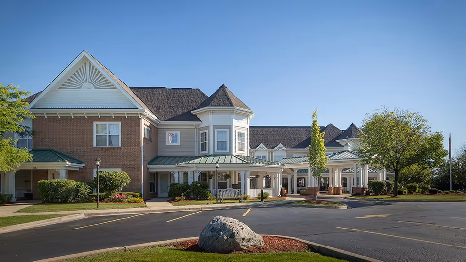 Exterior view of a senior living facility building with a combination of brick and light-colored siding, featuring a peaked roof and a covered entrance. The building is surrounded by neatly trimmed bushes, trees, and a parking lot with marked spaces under a clear blue sky.