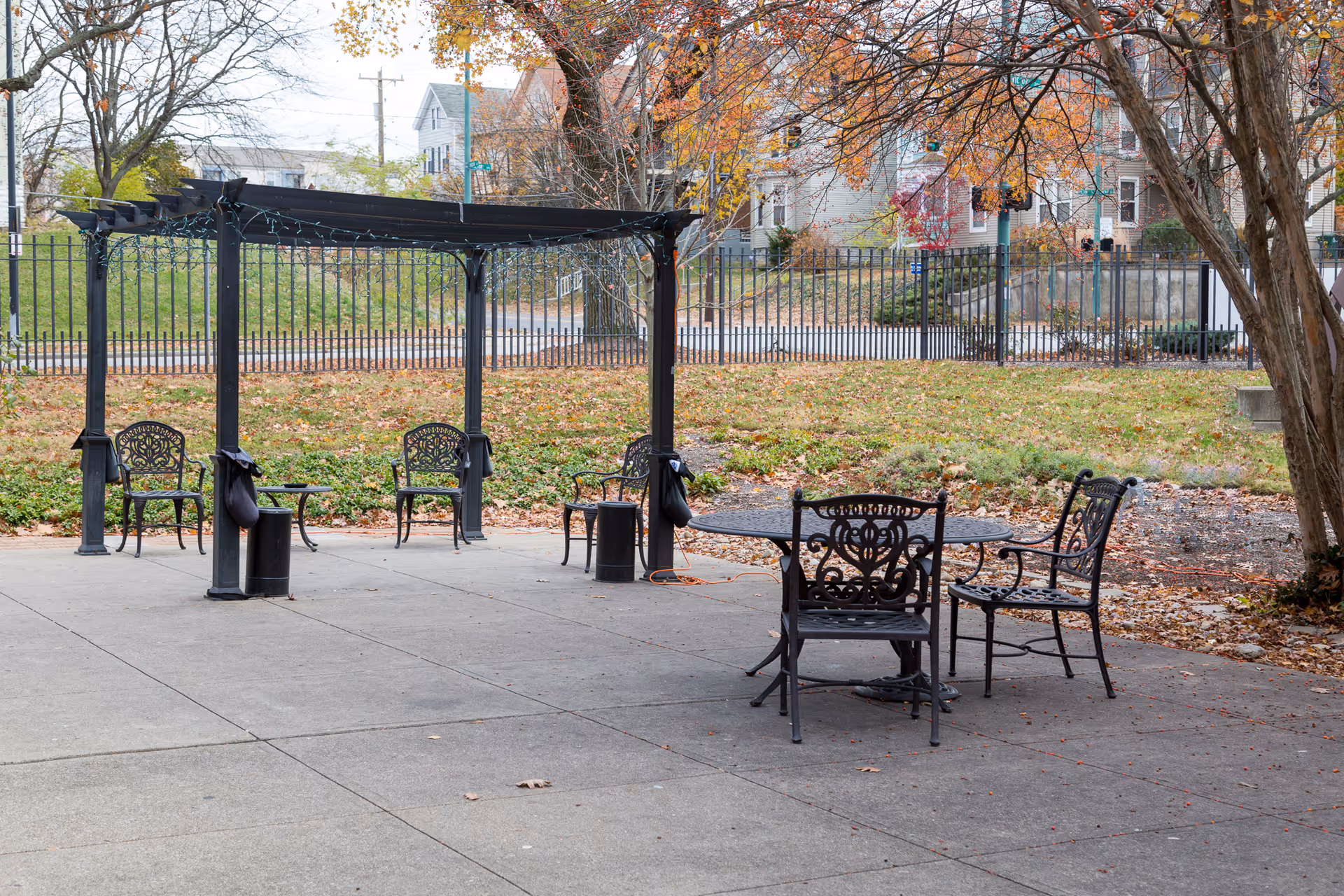 Outdoor patio area with black metal chairs and tables, a pergola structure with string lights, surrounded by trees with autumn leaves and a black metal fence in the background.