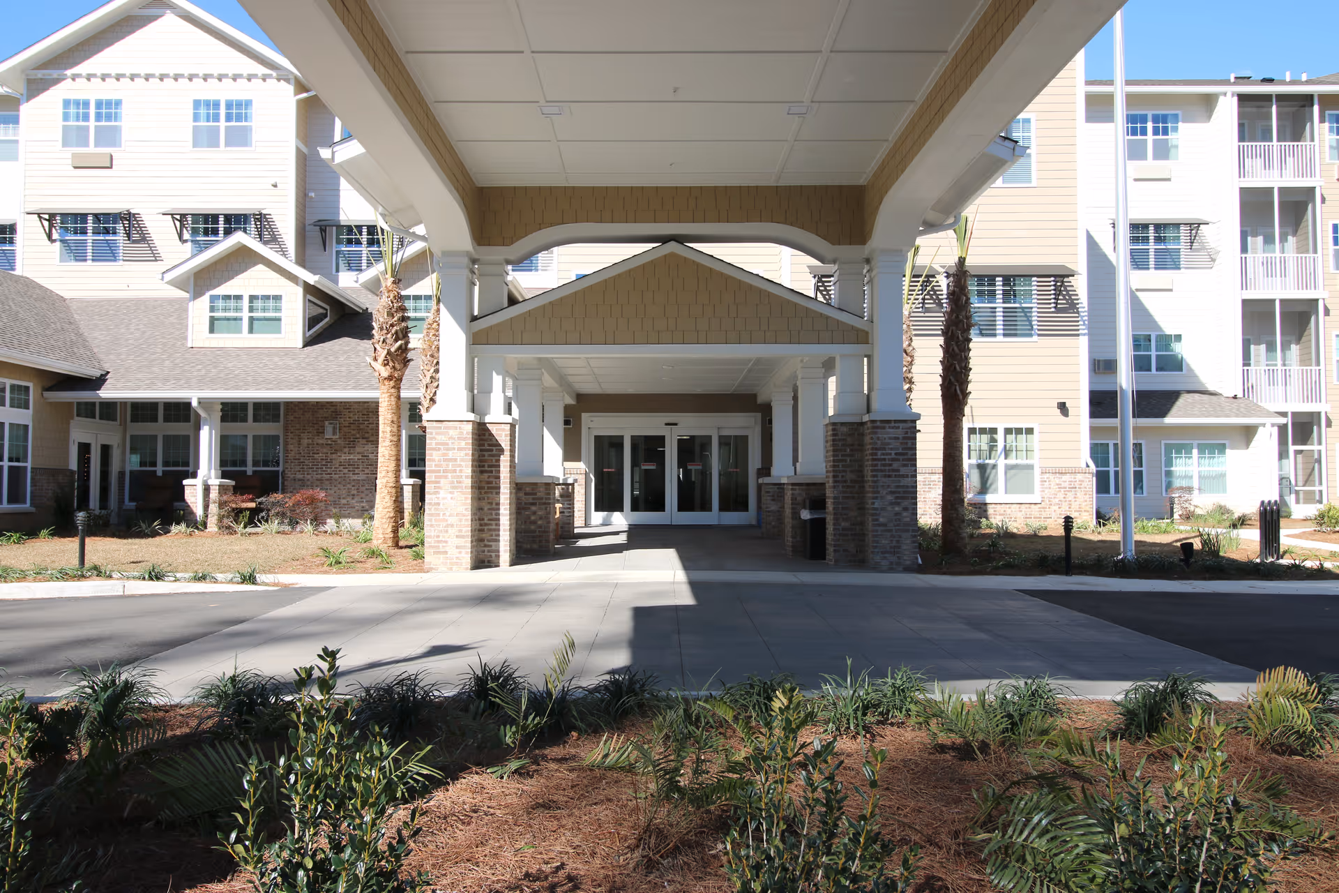 Entrance of a multi-story senior living facility with a covered driveway, brick pillars, and landscaped plants in front. The building has beige siding, multiple windows, and a clear blue sky above.