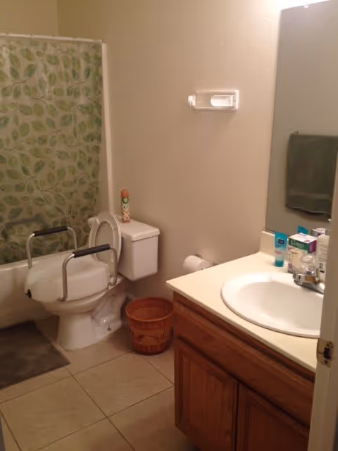 Small bathroom with a toilet fitted with a raised seat and safety rails, a sink vanity with toiletries, and a bathtub with a green leaf-patterned shower curtain.