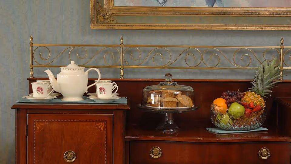 Ornate wooden sideboard displaying a teapot with cups, a glass cake dome of biscuits, and a bowl of assorted fruit beneath a framed painting.