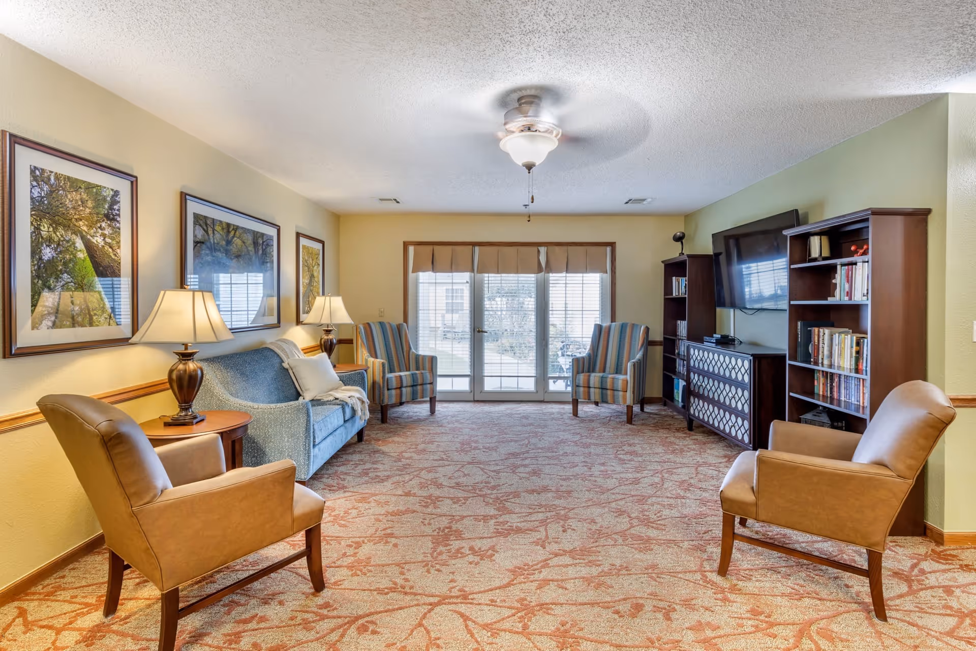 A well-lit living room with a patterned carpet, two brown leather armchairs, a blue upholstered sofa, two striped armchairs near glass doors, two wooden bookshelves, a flat-screen TV mounted on the wall, and framed nature photographs on the walls.