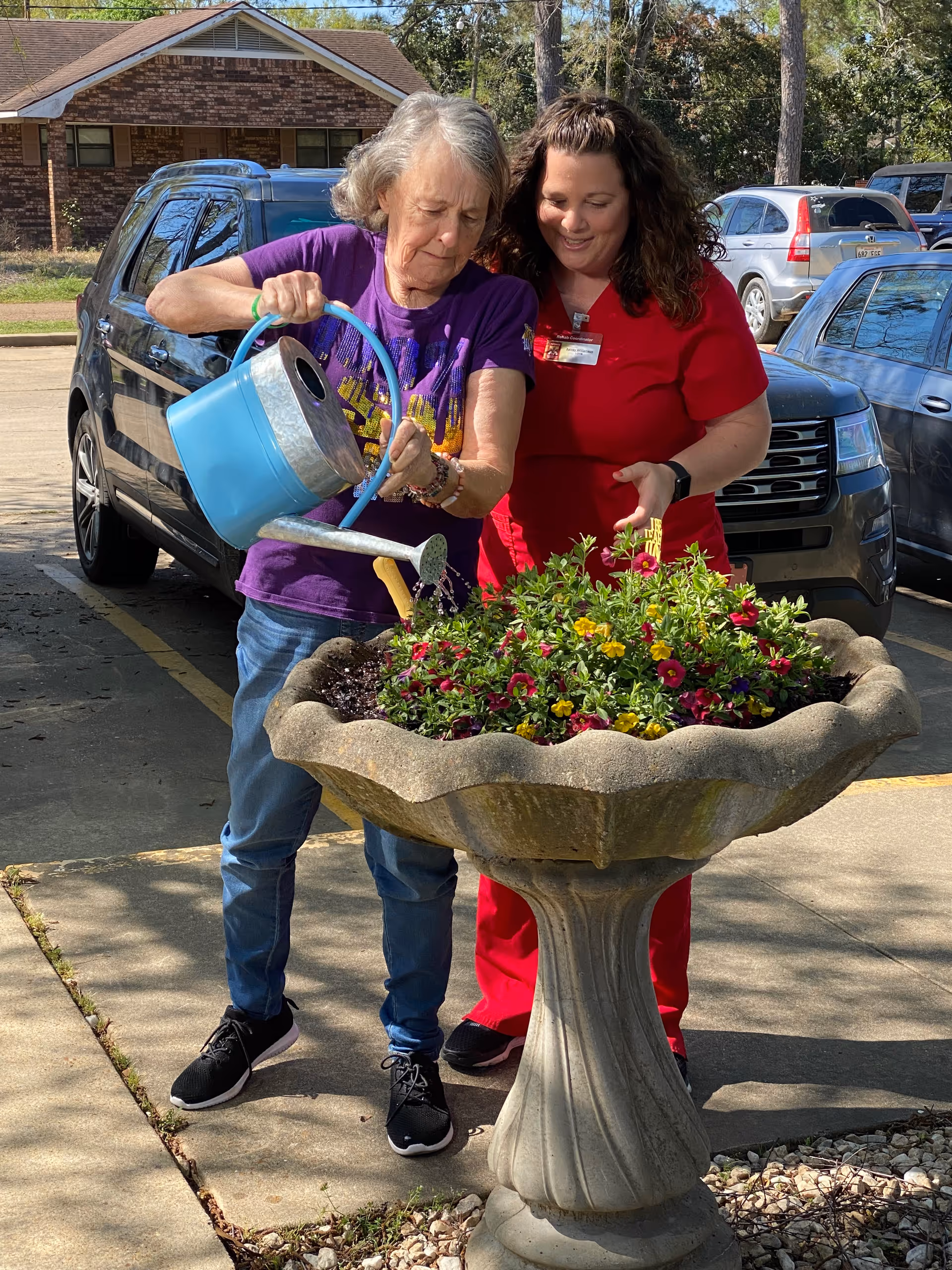 An elderly woman wearing a purple shirt and jeans waters flowers in a large stone planter with a blue watering can, while a woman in red scrubs stands beside her smiling and offering support. They are outdoors in a parking lot area with several cars and trees in the background.