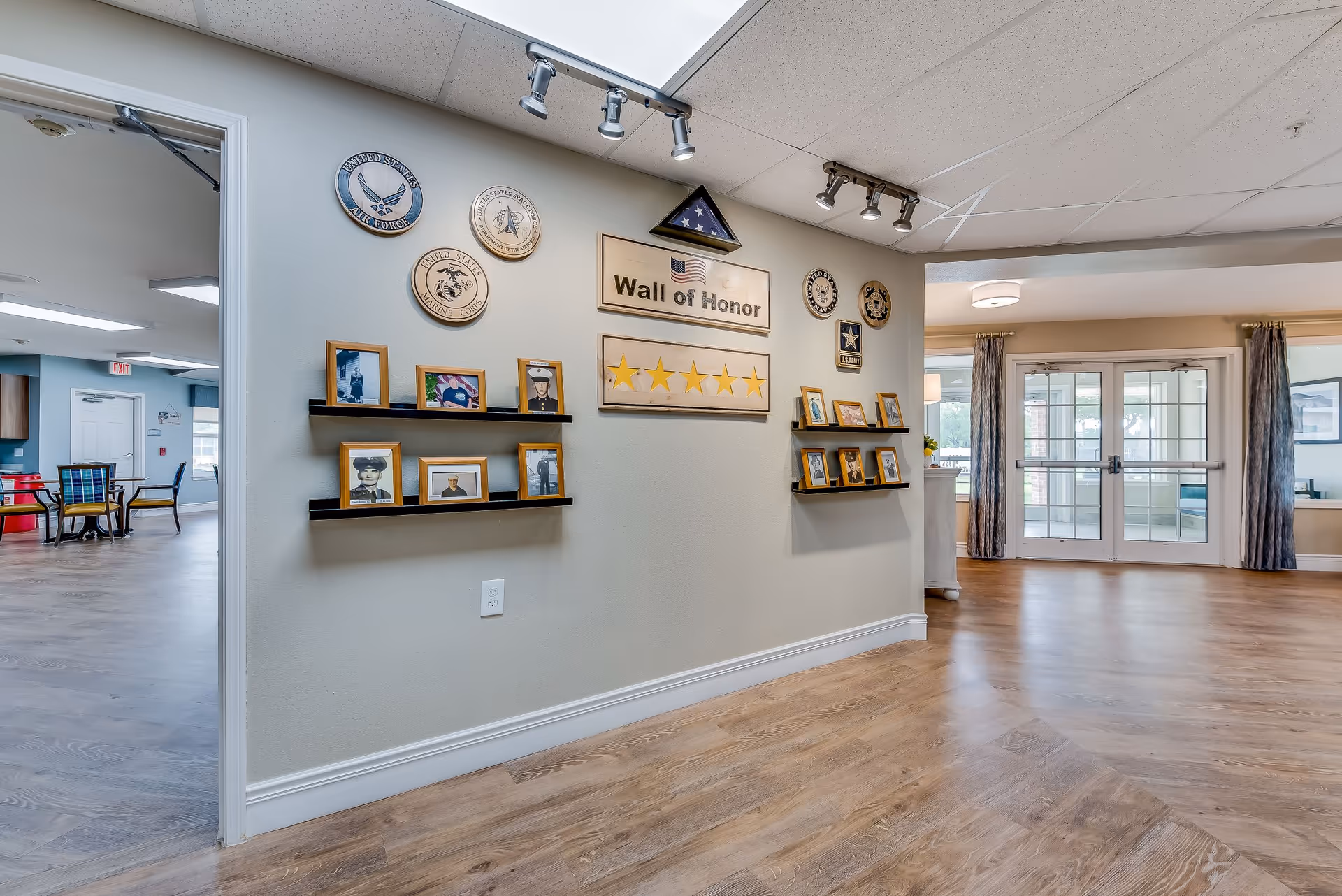 Interior view of a senior living facility hallway featuring a Wall of Honor display with framed photos, military insignias, and a folded American flag. The space has wood flooring, light-colored walls, and ceiling track lighting. In the background, there is a room with tables and chairs and glass double doors leading outside.