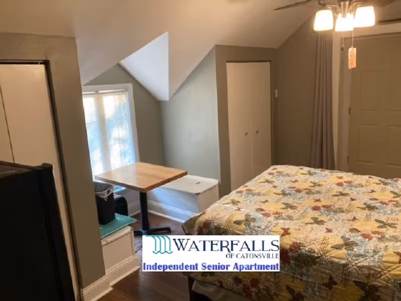 A small bedroom with a bed covered in a floral quilt, a wooden table near a window with blinds, a white storage bench, a closet with double doors, and a ceiling light fixture.