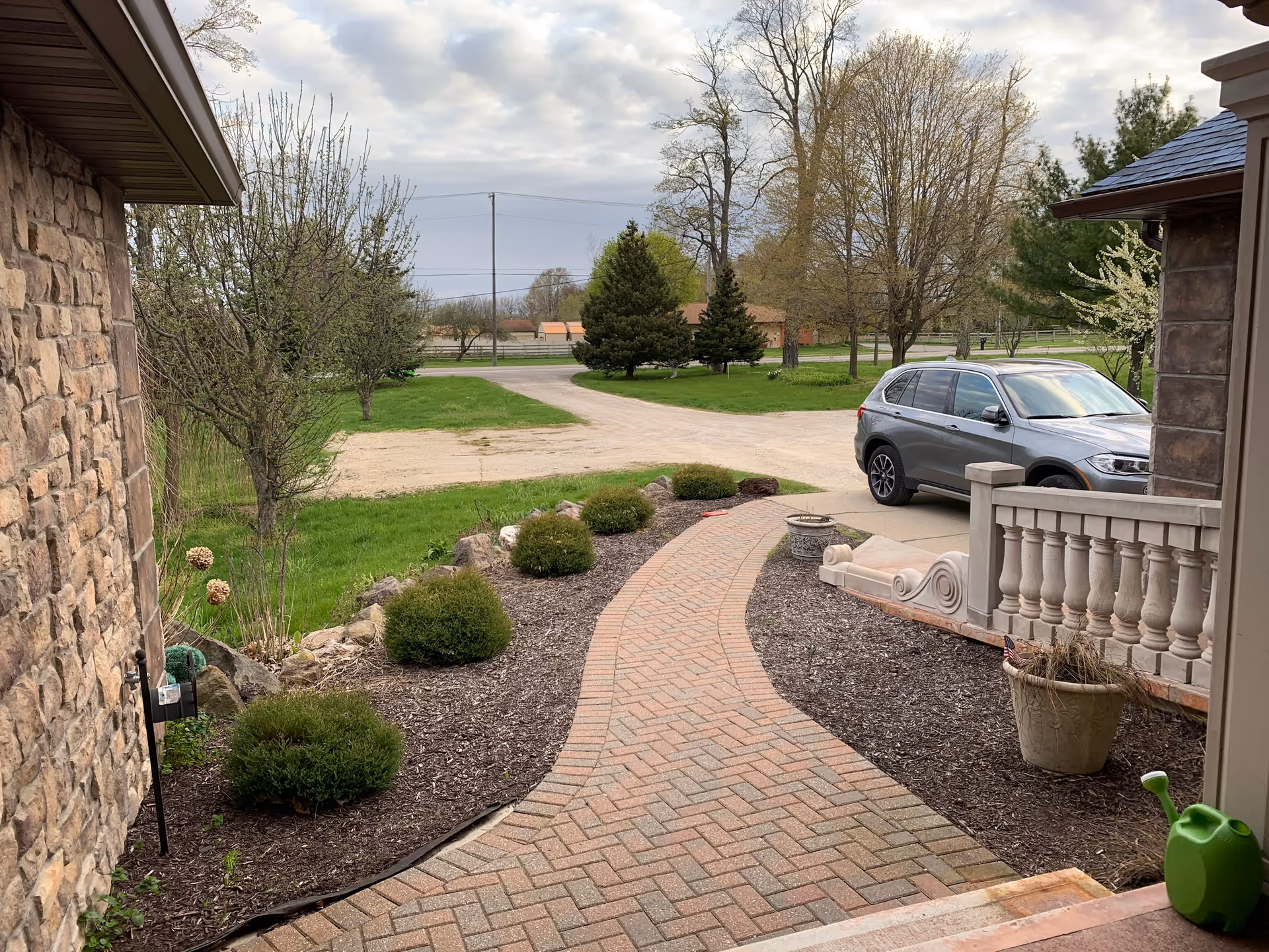 A paved brick walkway leading from a porch area with stone walls and decorative balusters, surrounded by mulch beds with small bushes. A gray SUV is parked on a driveway next to the porch. In the background, there are trees with budding leaves and a grassy area under a cloudy sky.