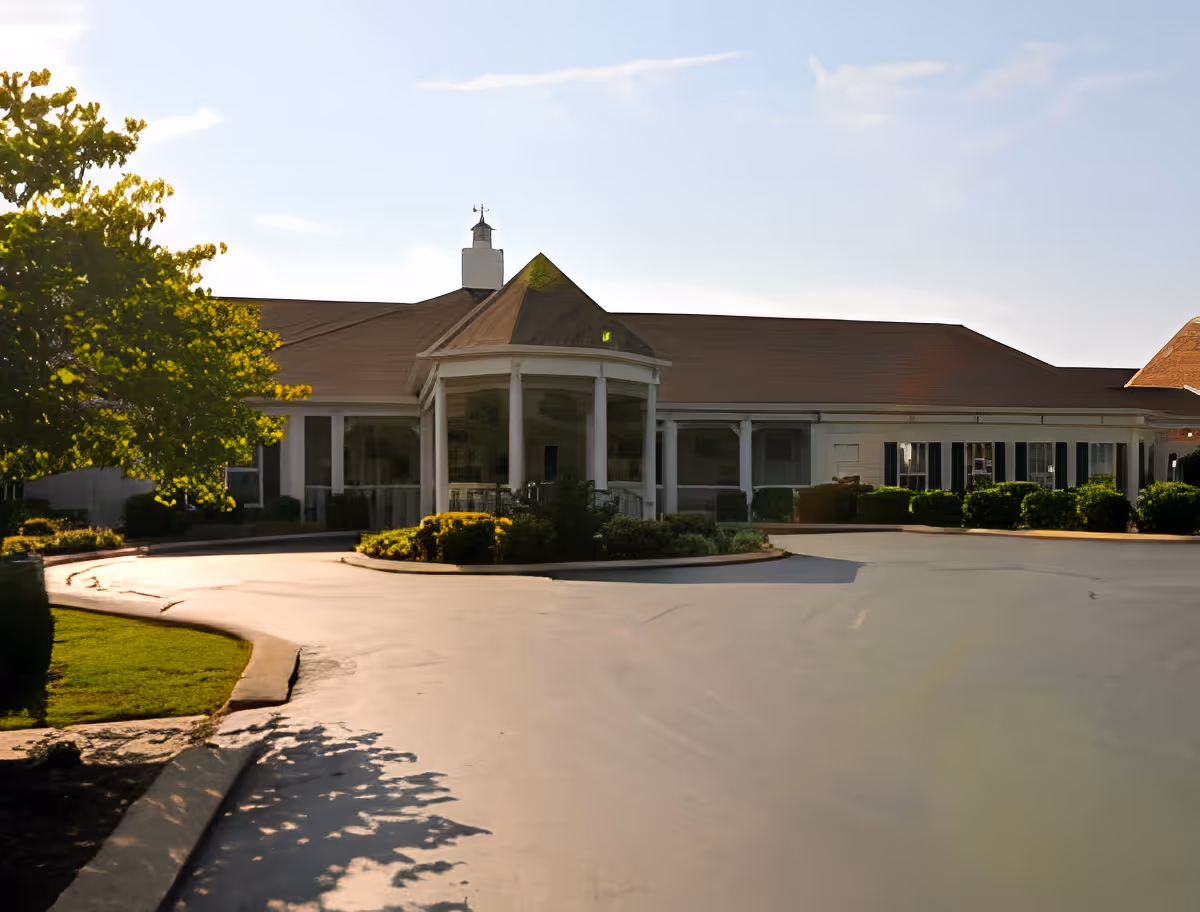Exterior view of a single-story senior living facility building with a covered entrance supported by white columns, surrounded by landscaped bushes and trees under a partly cloudy sky.