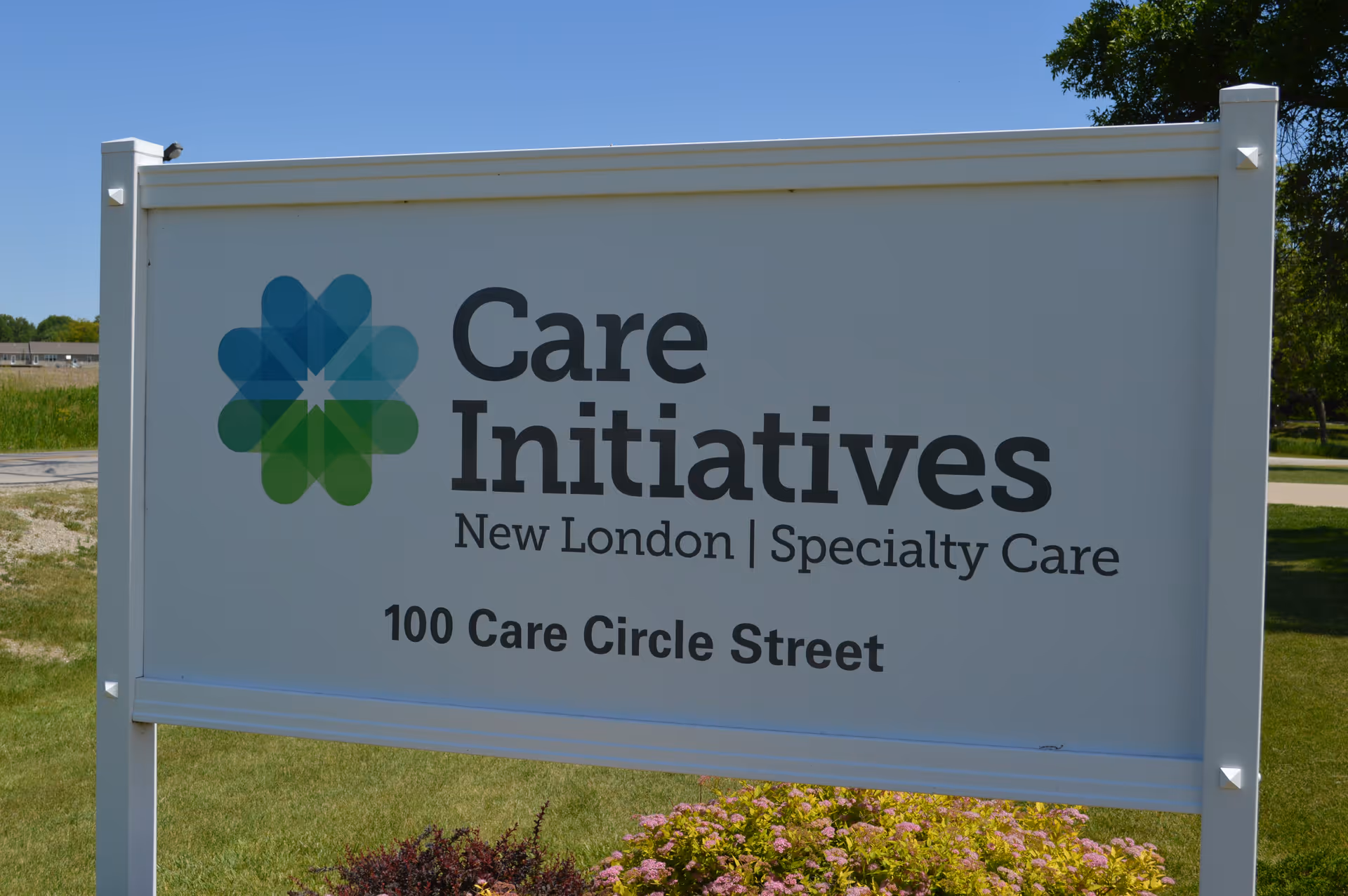 A white outdoor sign for Care Initiatives New London Specialty Care located at 100 Care Circle Street, with a blue and green geometric flower-like logo on the left side. The sign is set on a grassy area with some flowers and trees in the background under a clear blue sky.