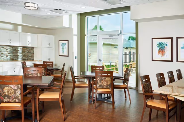 Dining room with several wooden tables and chairs, a kitchenette along one wall, and glass doors leading to an outdoor patio.