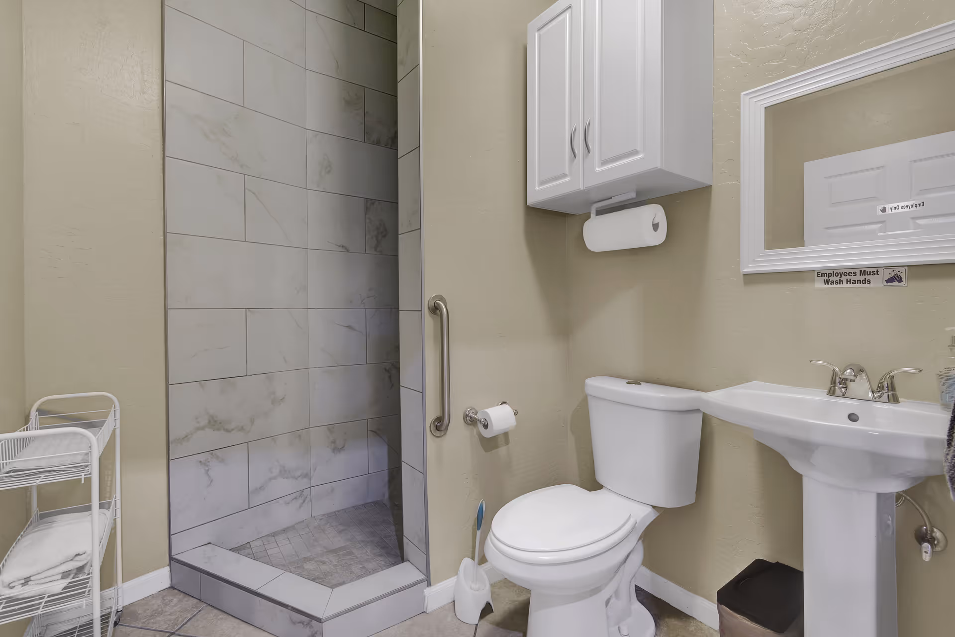 Small accessible bathroom with a tiled walk-in shower, toilet, pedestal sink, wall cabinet, and towel rack.
