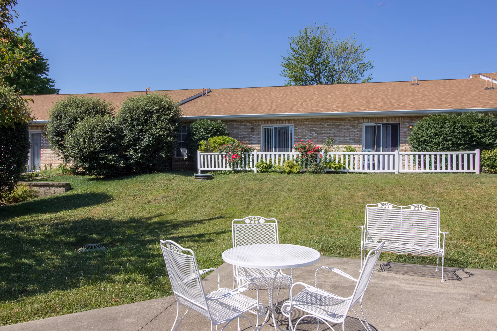 Outdoor patio with white metal table and chairs on a lawn in front of a single-story brick building with shrubs and a white railing.