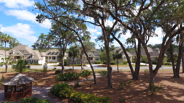 View of a senior living community with multiple beige residential buildings surrounded by trees and landscaped grounds under a partly cloudy blue sky. There is a small gazebo in the foreground and a paved walkway leading through the area.