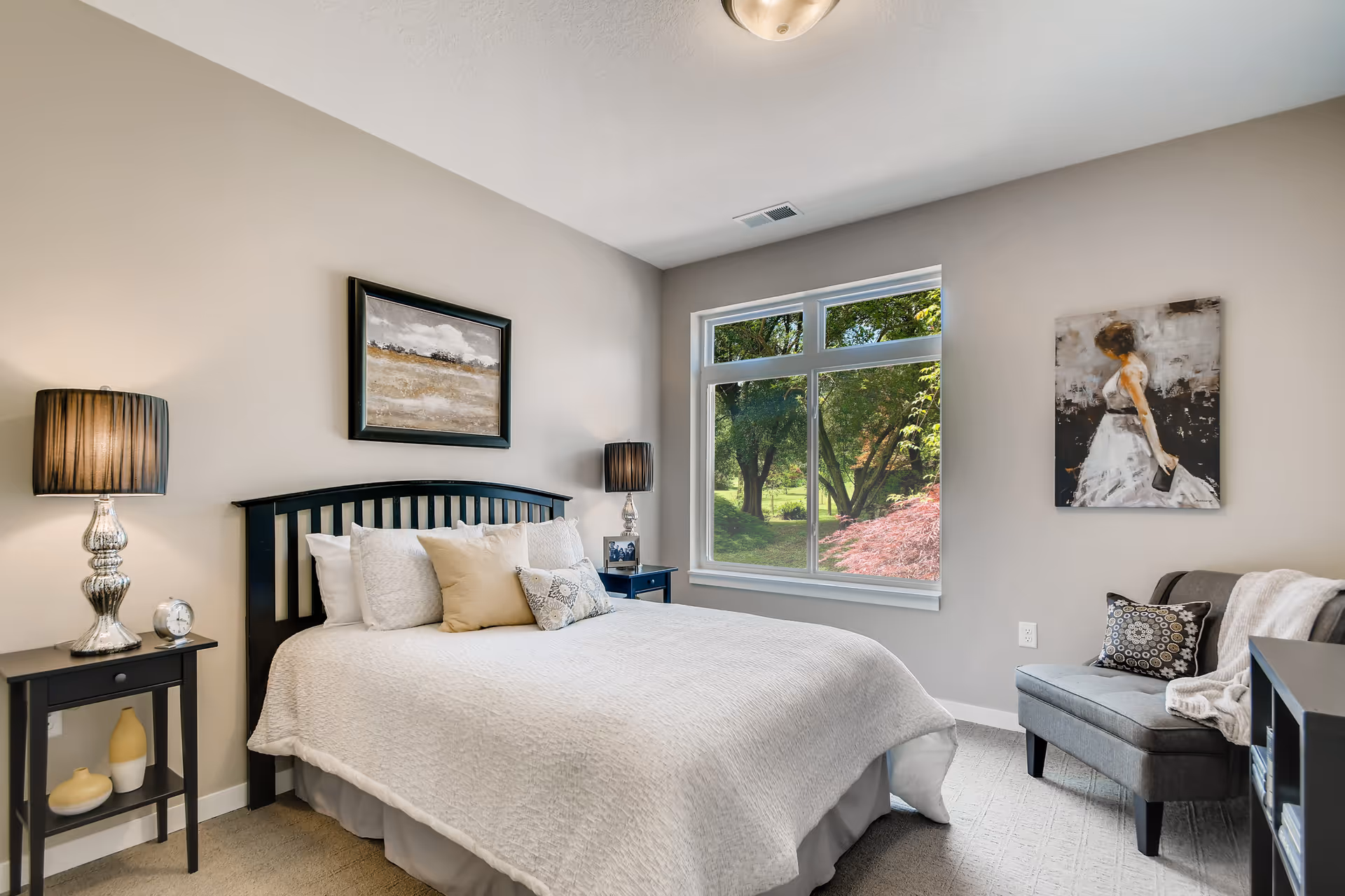 A cozy bedroom with a large bed covered in white bedding and multiple pillows. There are two matching bedside tables with silver lamps on each side of the bed. A framed landscape painting hangs above the headboard. A large window shows green trees and a garden outside. On the right side of the room, there is a gray upholstered chair with a decorative pillow and a throw blanket, and a painting of a woman in a white dress hangs on the wall above it.