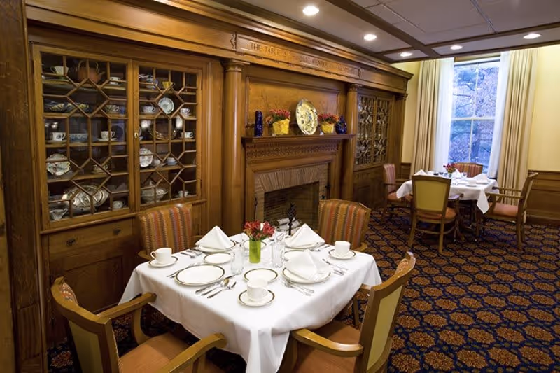 A cozy dining room with wooden paneling and built-in cabinets displaying china. Two tables are set with white tablecloths, plates, cups, silverware, and napkins, each with a small vase of red flowers. A fireplace with decorative plates and vases sits between the cabinets, and a large window with cream curtains lets in natural light.