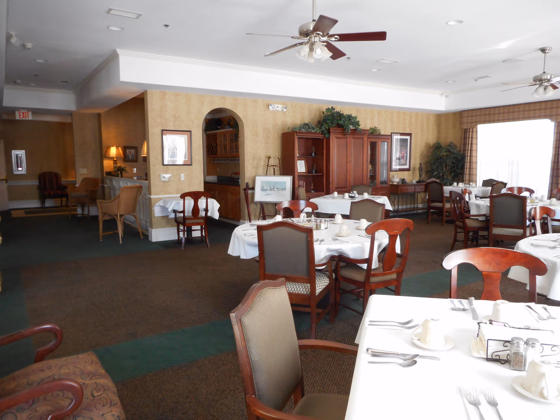 A spacious dining room with multiple round tables covered with white tablecloths, set with cups, plates, and silverware. The room features wooden chairs with cushioned seats, a ceiling fan, and a large window with curtains allowing natural light. There is a wooden cabinet with decorative plants and framed artwork on the walls.