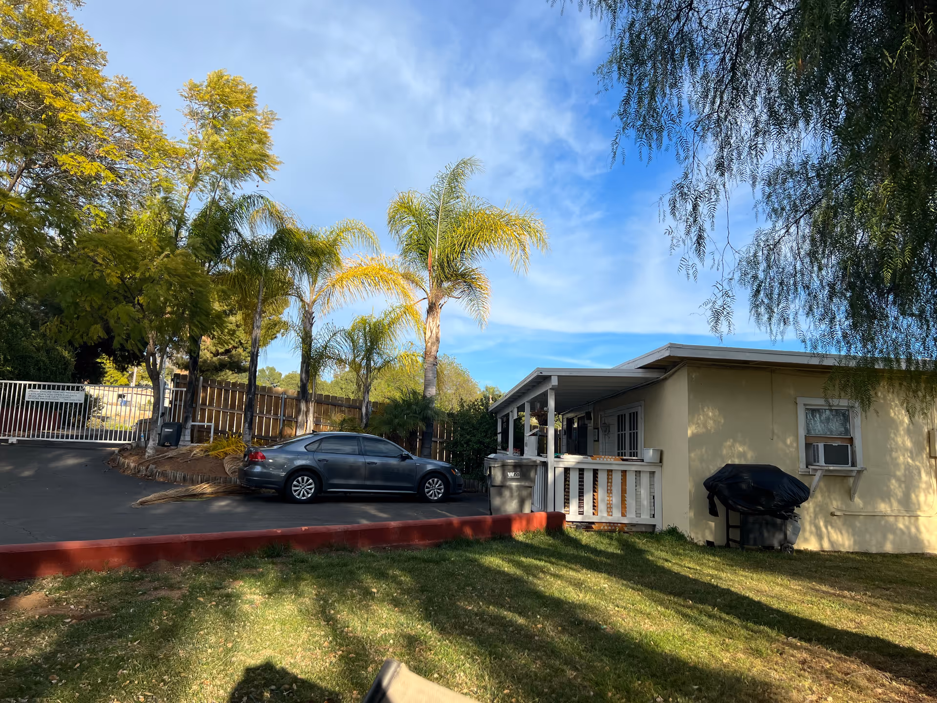Single-story beige building with a covered porch, parked car, palm trees, and a gated driveway under a blue sky.