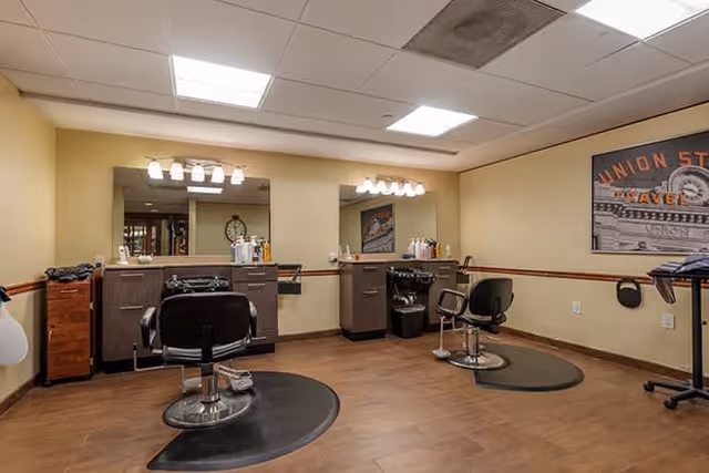 Interior view of a salon area in a senior living facility with two black salon chairs in front of large mirrors mounted on beige walls. The room has wooden flooring, overhead fluorescent lighting, and various hair care products on the counters. A framed picture is visible on the right wall.