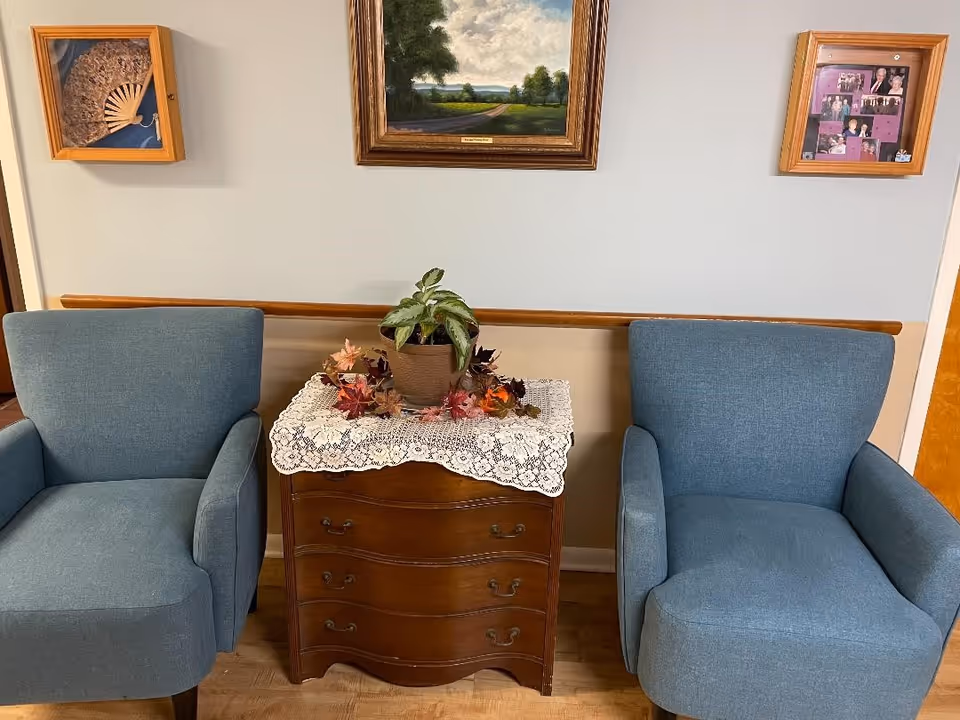 Two blue armchairs flank a small wooden chest topped with a potted plant and a lace doily beneath framed wall art.