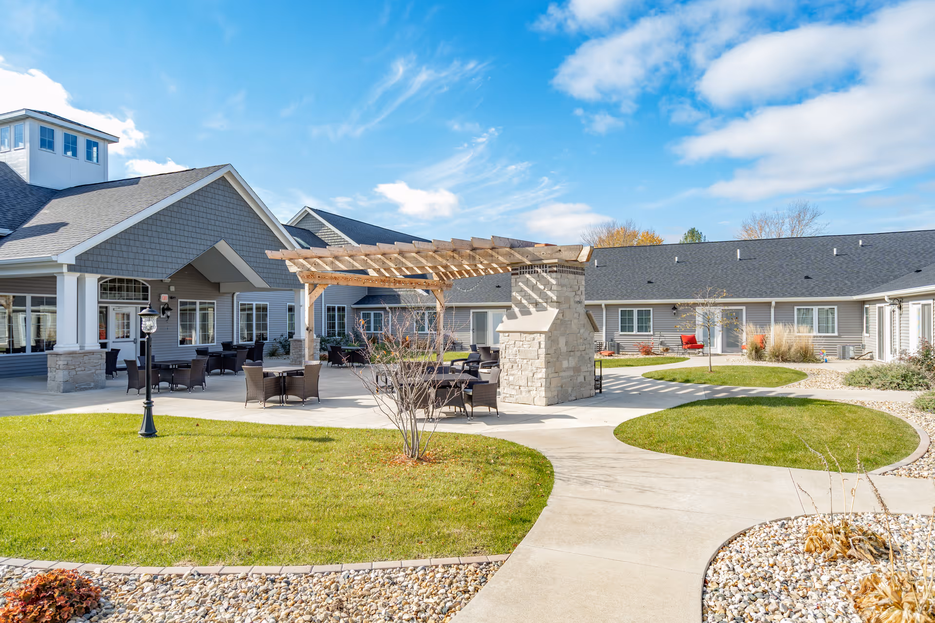 Outdoor courtyard area of Villas of Holly Brook Assisted Living & Memory Care in Pekin, IL, featuring a paved walkway, green lawn, a pergola with stone pillars, outdoor seating with tables and chairs, and a building with gray siding and white trim under a partly cloudy blue sky.