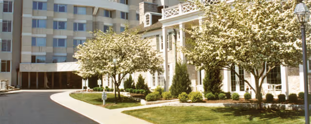 Front entrance and facade of the Wesley Glen senior living building with a curved driveway, landscaped lawn, and flowering trees.