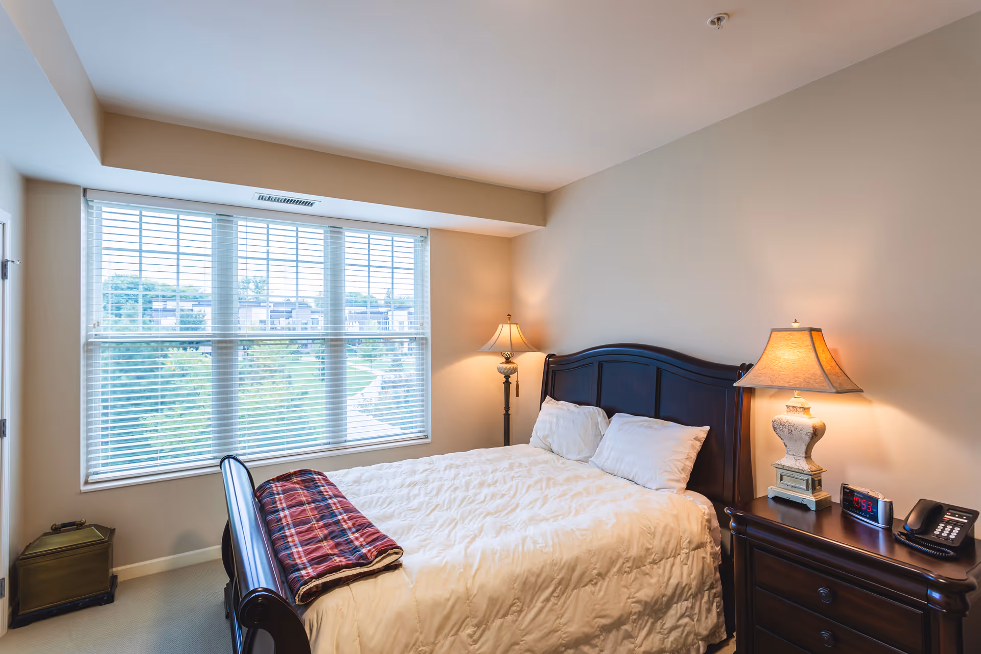 A cozy bedroom with a large window covered by white blinds, a bed with white bedding and two pillows, a red plaid blanket folded at the foot of the bed, two bedside tables each with a lamp, a digital alarm clock, and a telephone.