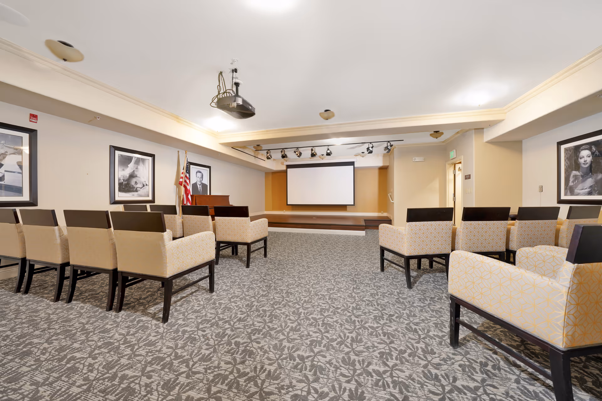 Carpeted meeting room with rows of upholstered chairs facing a small stage and projection screen.