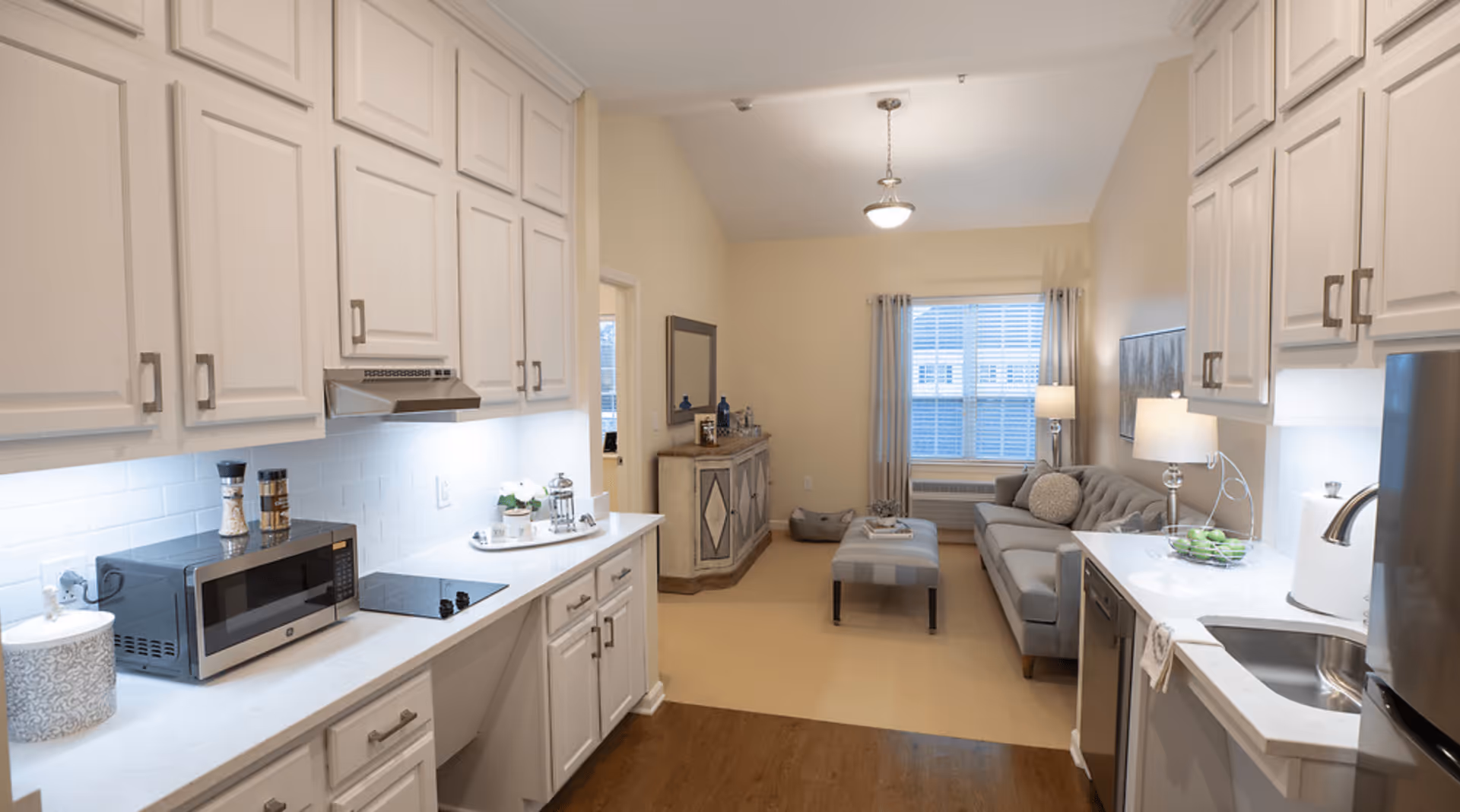 A bright and modern kitchen and living room area in a senior living facility. The kitchen features white cabinetry, a microwave, stovetop, and a sink with a stainless steel faucet. The living room has a gray sofa, a striped ottoman, a floor lamp, a window with curtains, and a sideboard with decorative items.