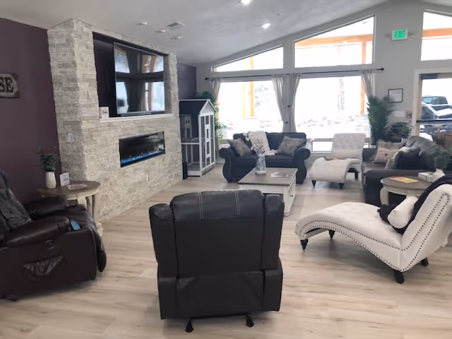 A spacious living room with light wood flooring, featuring a stone fireplace with a mounted TV above it. The room contains multiple seating options including a dark leather recliner, a white chaise lounge, a gray sofa, and a cushioned armchair. Large windows with sheer curtains allow natural light to fill the space, and there are small tables and decorative plants around the room.