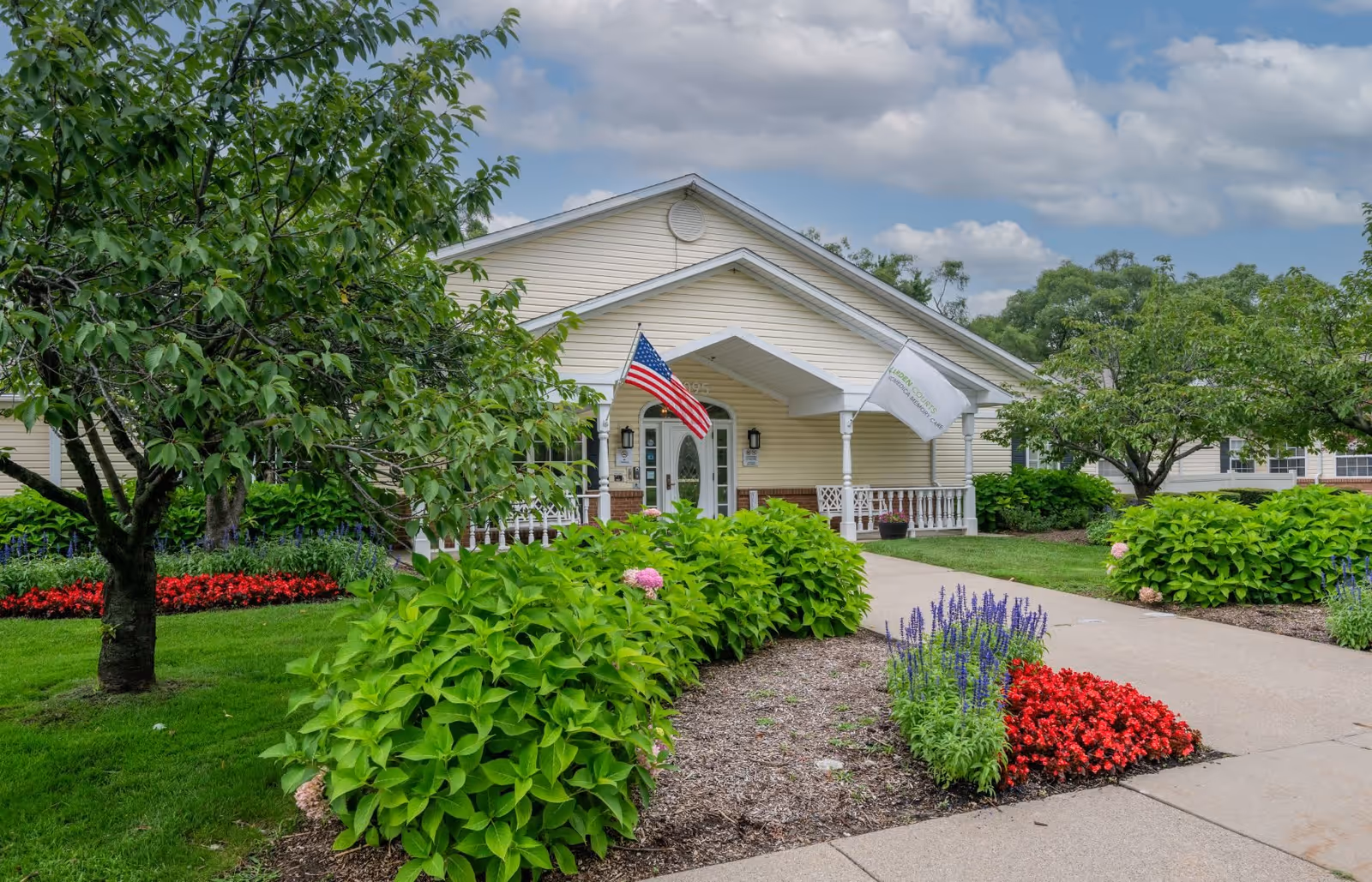 Front exterior view of a single-story building with beige siding and a white porch. The entrance is adorned with an American flag and another flag. The building is surrounded by well-maintained landscaping including green bushes, colorful flowers, and trees under a partly cloudy sky.
