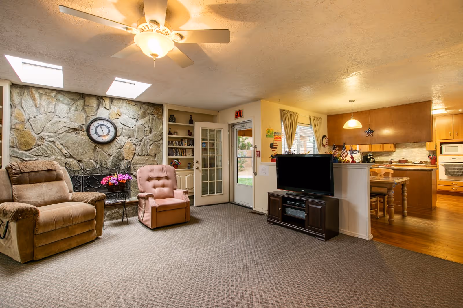 A cozy living room area with two recliner chairs, a stone accent wall with a clock, a ceiling fan with light, and a TV on a stand. The room opens into a kitchen and dining area with wooden cabinets and a dining table with chairs. There is a glass door leading outside and windows with curtains letting in natural light.