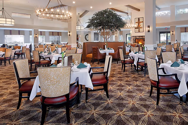A spacious and elegant dining room with multiple tables covered in white tablecloths, each set with green napkins and glassware. The room features patterned carpet, high ceilings with modern chandeliers, large windows allowing natural light, a large indoor tree, and a cozy fireplace in the background.