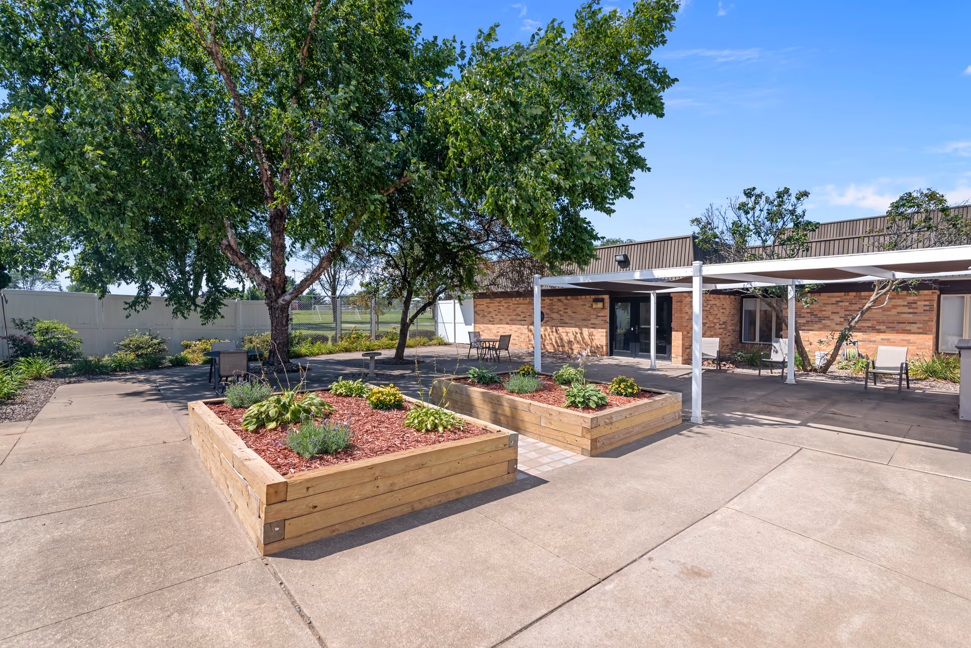 Outdoor patio area at Edenbrook Platteville with raised wooden garden beds containing plants and flowers, large trees providing shade, several chairs and tables, and a brick building in the background under a clear blue sky.