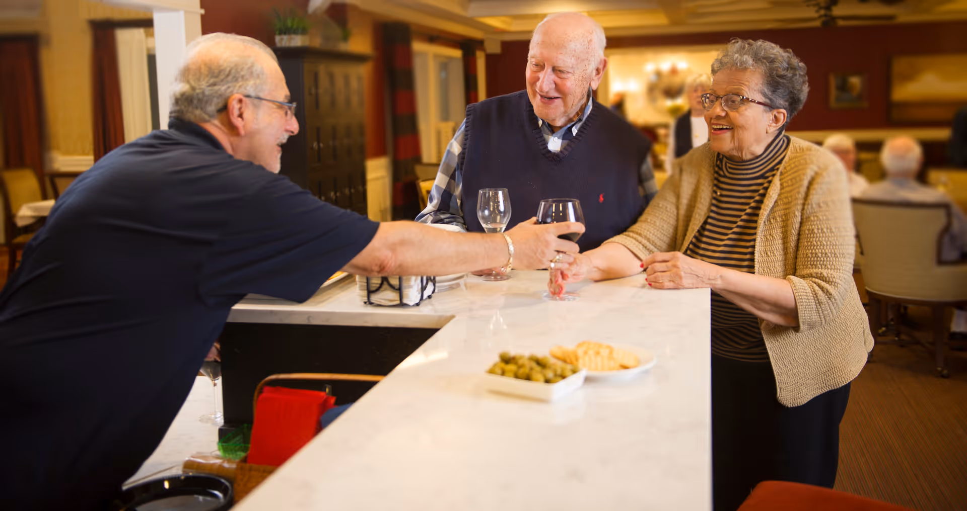 Three older adults chatting and enjoying wine at a light marble bar counter with a small plate of olives and crackers.
