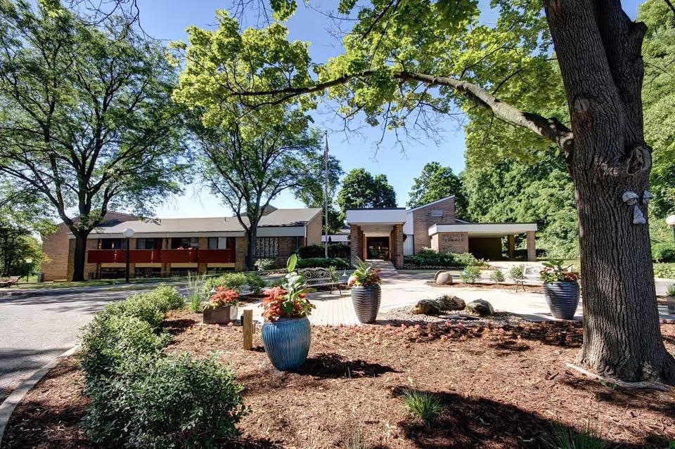 Exterior view of Hillside Terrace Senior Living facility showing a brick building surrounded by trees and landscaped garden beds with large planters and benches under a clear blue sky.