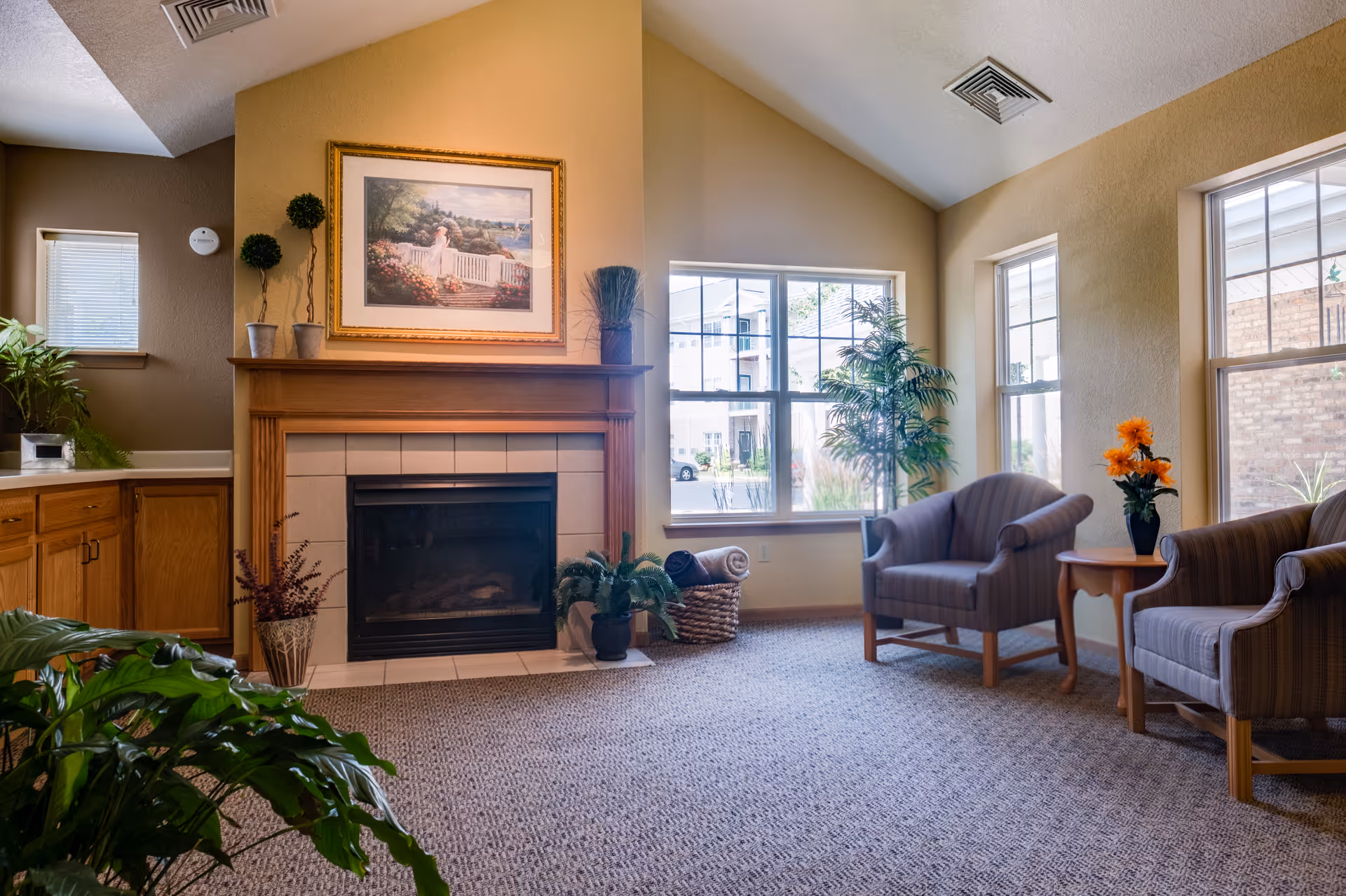 A cozy living room area with a fireplace centered on a beige wall. Above the fireplace is a framed painting of a woman in a garden. To the left of the fireplace are wooden cabinets and a small window. To the right, there are three large windows letting in natural light, two striped armchairs, a small wooden side table with a vase of orange flowers, and several potted plants around the room. The carpet is a neutral tone, and the ceiling is vaulted with recessed lighting.